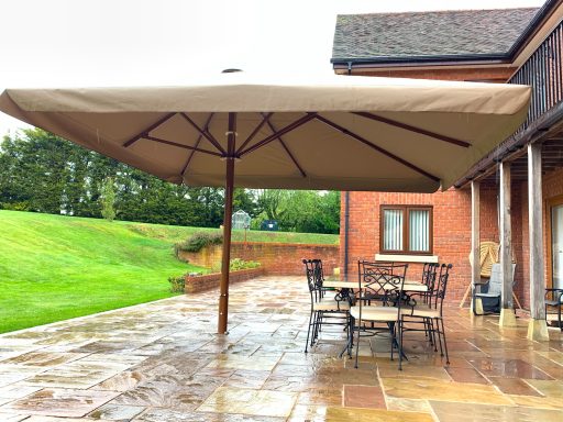 Large beige umbrella over a wooden table and chairs on a stone patio, amid greenery.