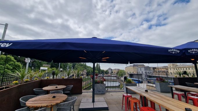 Outdoor café with red and wooden tables under large blue umbrellas by a waterway.