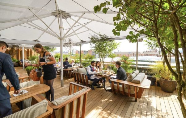 Outdoor dining area with wooden tables, greenery, and patrons enjoying their meals.