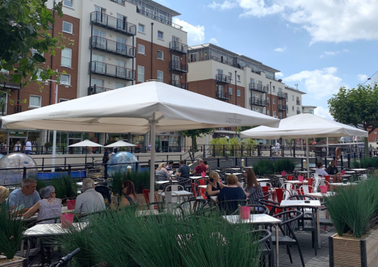 Outdoor dining area with tables under white umbrellas and patrons enjoying meals.