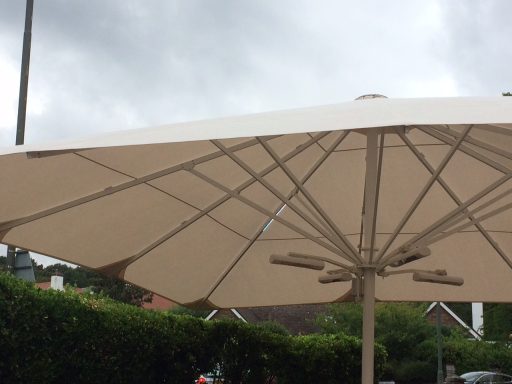 A large beige parasol against a cloudy sky, with a green hedge in the background.