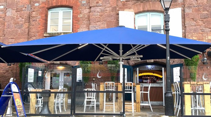 Exterior of a restaurant with blue umbrellas and outdoor seating on a brick façade.