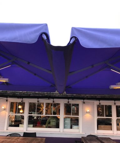 Purple awning above a restaurant with outdoor seating and large windows.