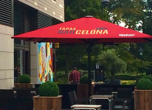 Red umbrella with "Barcelona" signage over outdoor seating surrounded by greenery.