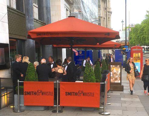 Crowd gathered at an outdoor café with orange umbrellas and plant decorations.