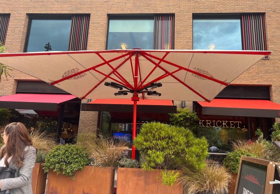 A red parasol shades a cafe entrance with greenery in front and a person in grey attire.