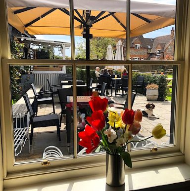 A sunny café patio view with flowers in a vase and people seated outdoors.