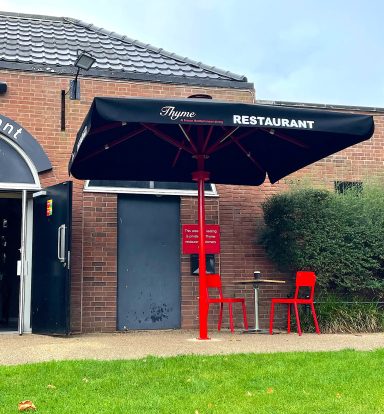 Black umbrella with "RESTAURANT" text over red chairs and a small table outside a brick building.