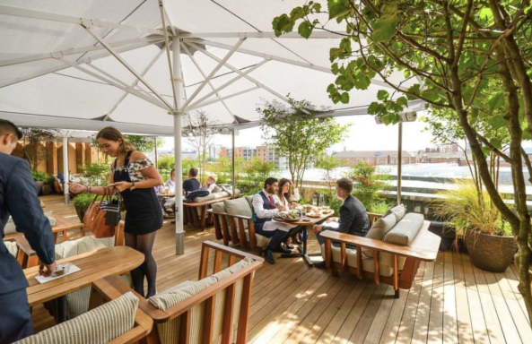 Outdoor dining area with wooden furniture, plants, and customers enjoying food and drinks.