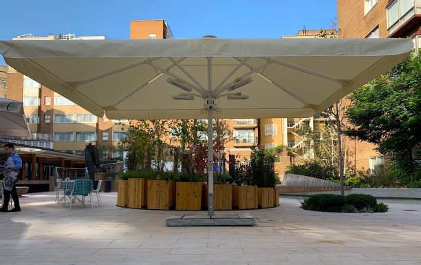 Large beige umbrella shading a circular planter with greenery in an urban setting.