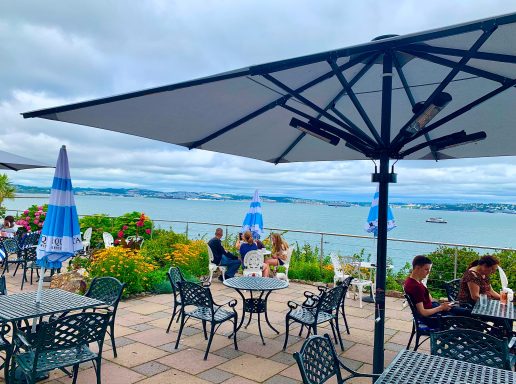 Outdoor café setting with tables, umbrellas, and views of a body of water and cloudy sky.