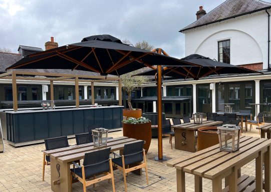 Outdoor seating area with wooden tables, chairs, and large umbrellas under a cloudy sky.