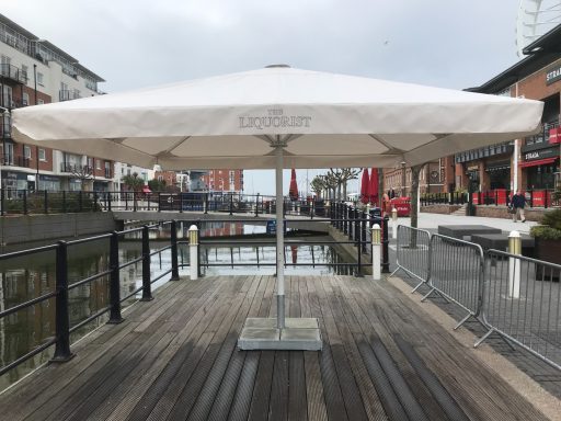 Large white parasol on a wooden deck by a canal, with buildings in the background.