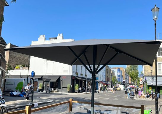 A large grey umbrella on a sunny street with shops and people in the background.