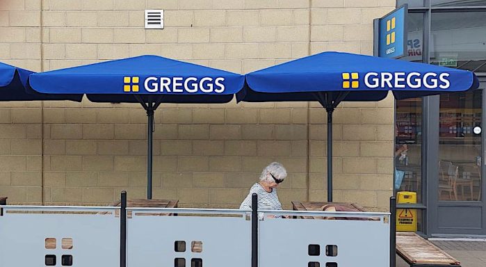 Outdoor seating area with blue umbrellas branded with "Greggs." A person is sitting at a table.