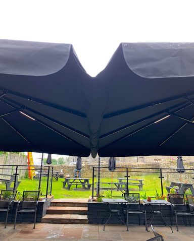 View of a black awning with a rain cover, outdoor seating area and greenery in the background.