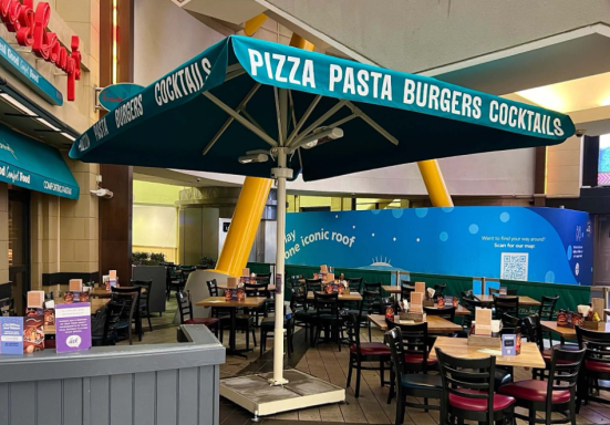 Umbrella-shaded dining area with tables at a casual restaurant in a food court.