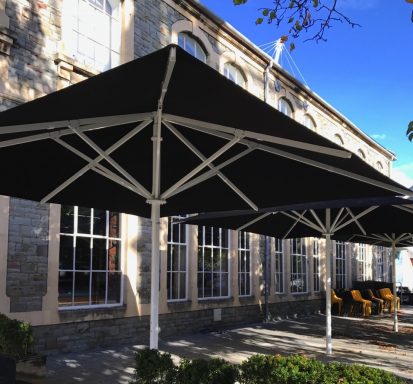 Black outdoor umbrellas provide shaded seating outside a stone building.