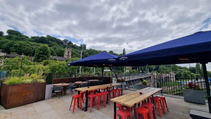 Outdoor cafe with red stools and blue umbrellas, surrounded by greenery and cloudy sky.