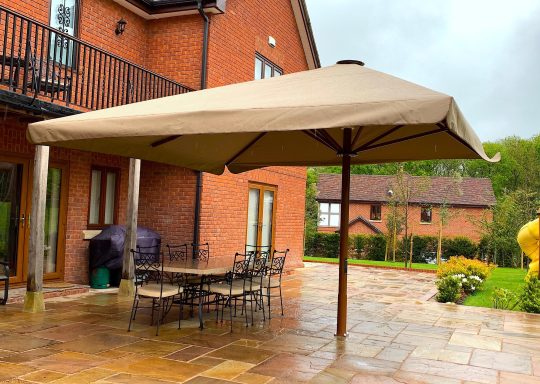 A large beige garden parasol over a table and chairs, beside a red brick house.