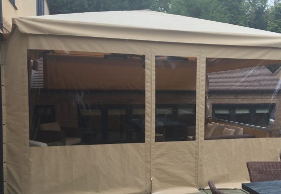 A tan outdoor gazebo with transparent windows, set in a garden.