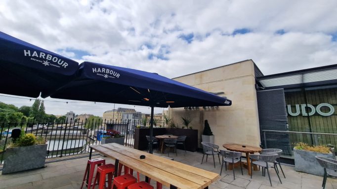 Outdoor dining area with tables, chairs, and large umbrellas beside a canal.