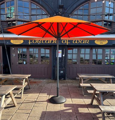 Outdoor seating area with wooden tables beneath an orange parasol and a pub in the background.