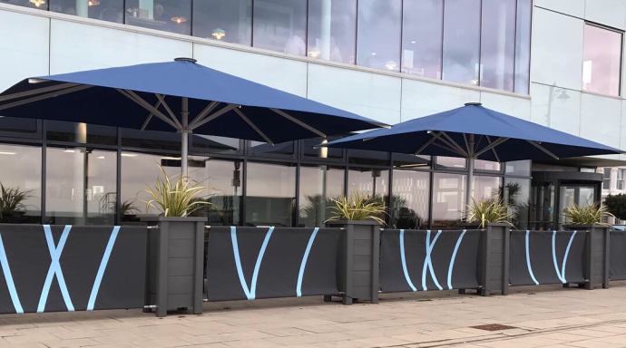 Outdoor seating area with blue umbrellas and planters along a building's facade.